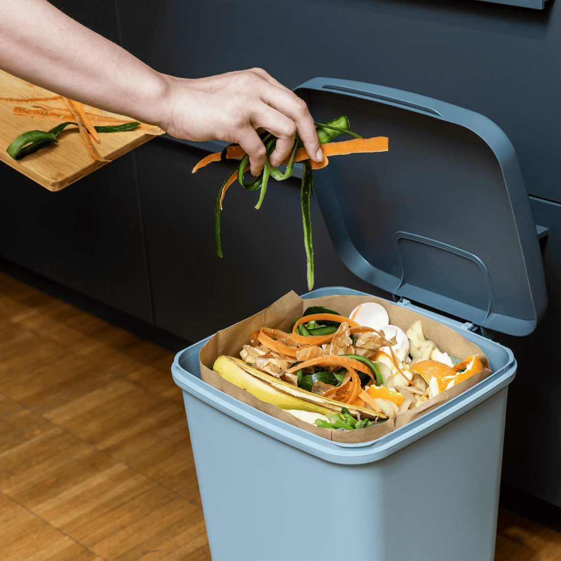 Hand gibt Gemüsereste in einen Papier-Biomüllbeutel mit Blockboden im blauen Bioeimer