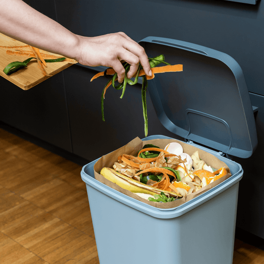 Hand gibt Gemüsereste in einen Papier-Biomüllbeutel mit Blockboden im blauen Bioeimer